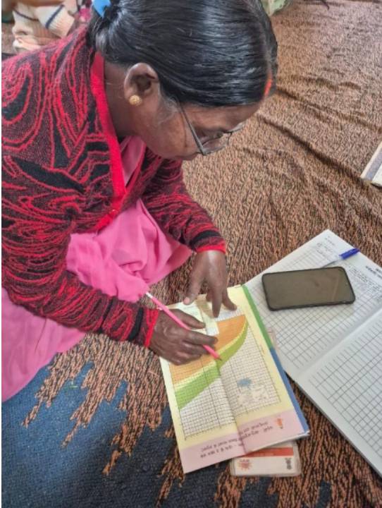 A woman sitting on the floor, wearing a red sweater and pink sari, focused on drawing a chart in a notebook with a pink pencil, while a smartphone and another notebook are visible nearby.