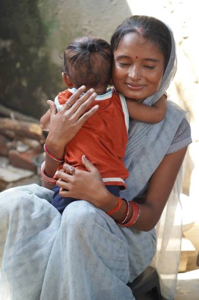 A woman in traditional attire embracing a child from behind, both showing warmth and affection, with a rural background.