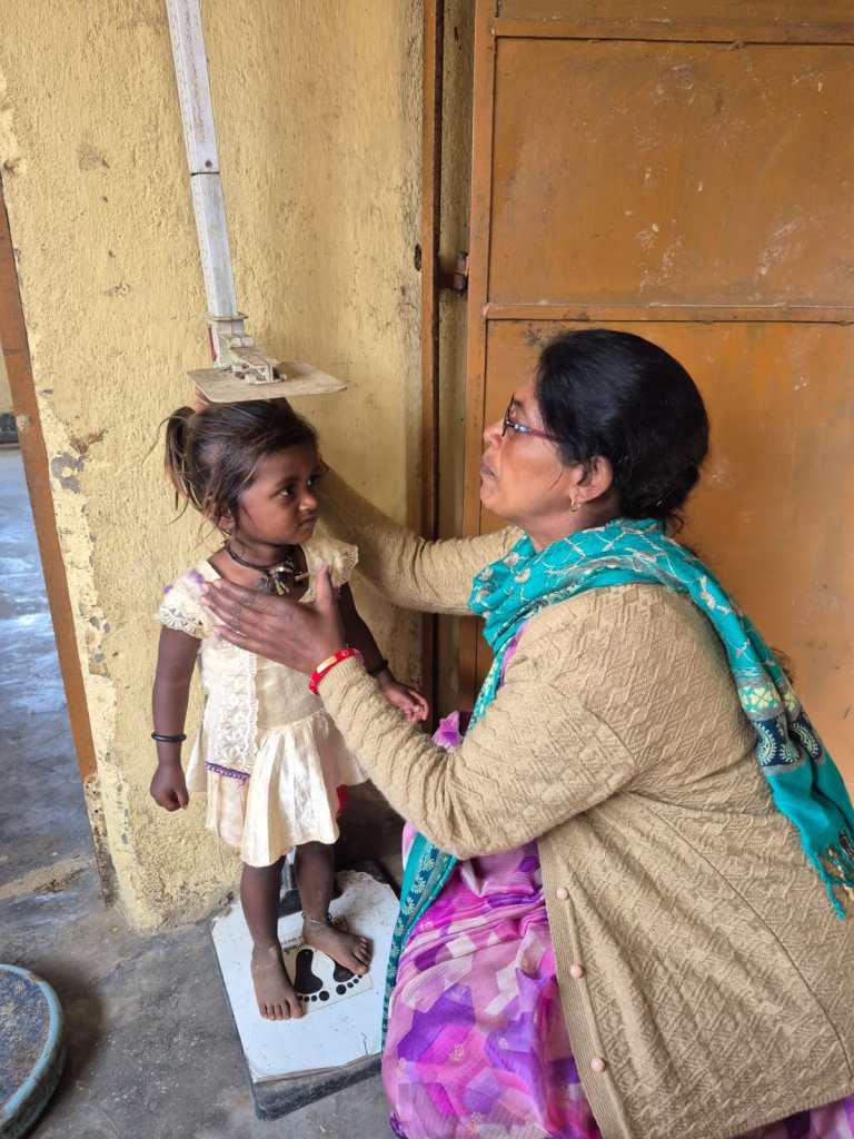 A woman measuring the height of a young girl standing on a weighing scale, in a room with a rustic wall.