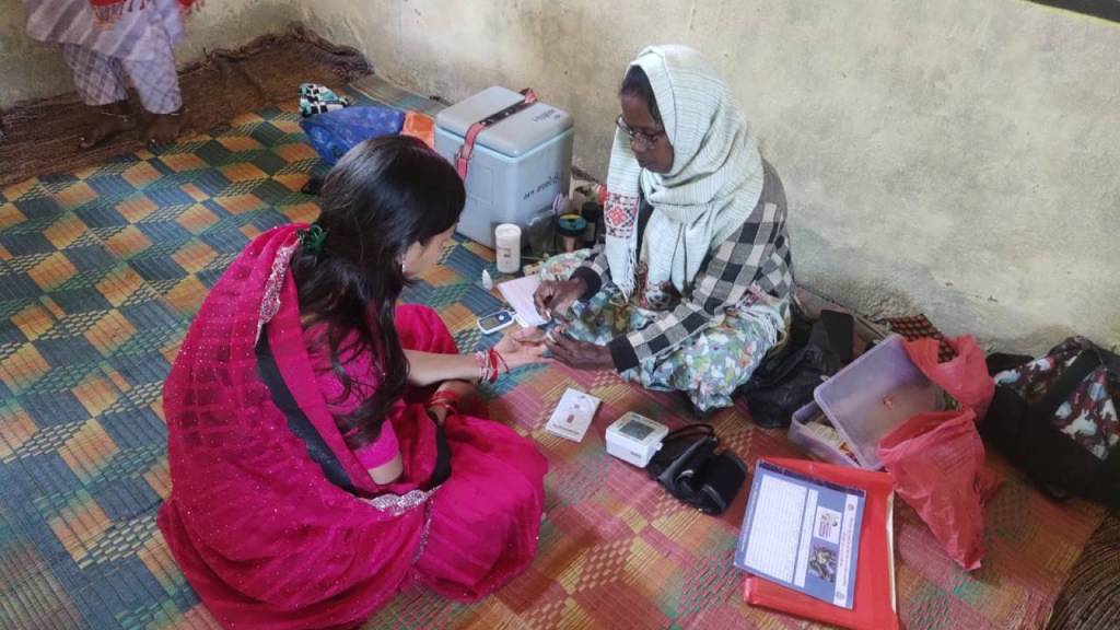 A woman in a traditional red sari sits on the floor engaged in a conversation with another woman wearing a scarf and patterned clothing. They are surrounded by various items including documents and containers on a colorful woven mat.