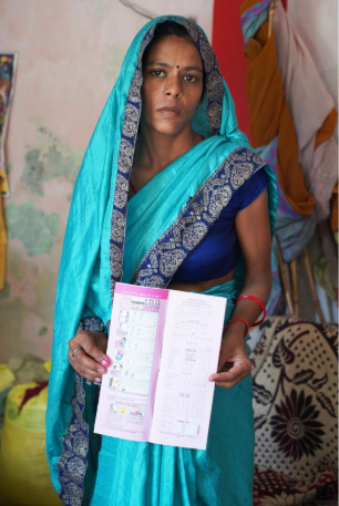A woman wearing a turquoise saree holds a pink document, looking directly at the camera. The background features colorful fabrics and bags.