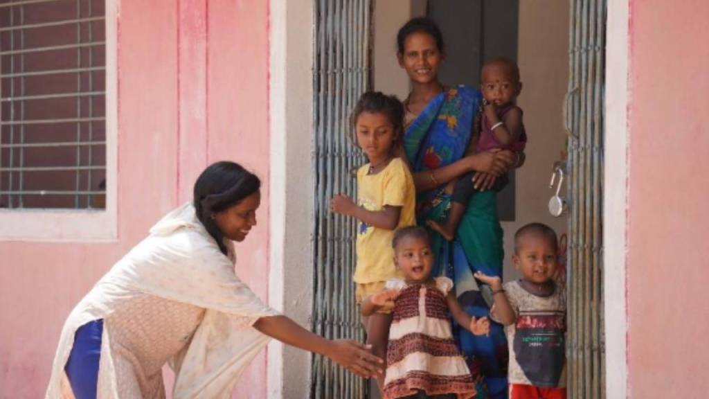 A woman in a white scarf interacts with young children outside a colorful building, while another woman holds a baby in her arms, surrounded by four cheerful children.