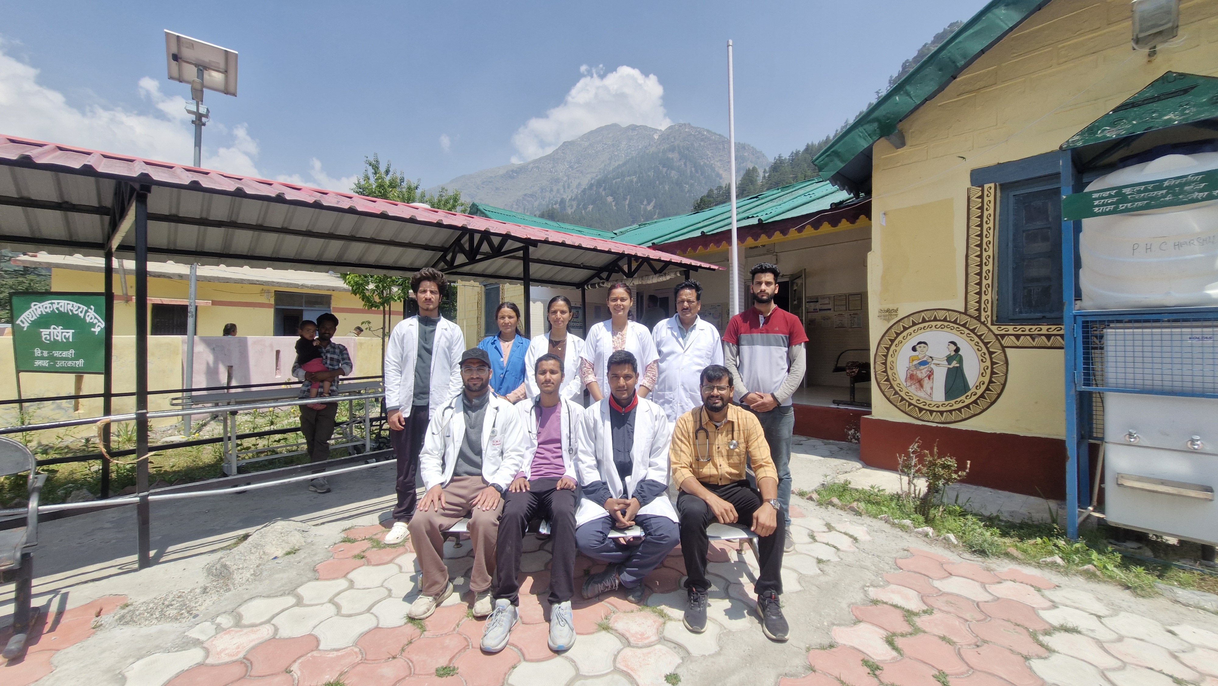 Group photo of healthcare workers in white coats outside a health center in a mountainous area, with a building and solar panel in the background.