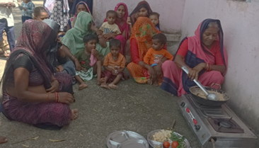 A group of women and children gathered outdoors, watching a woman cooking on a portable stove, with vegetables and ingredients laid out nearby.