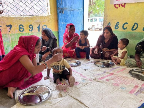 A group of women and children sitting on a mat in a room, enjoying a meal together. The women are feeding the children, and colorful decorations are visible on the walls.