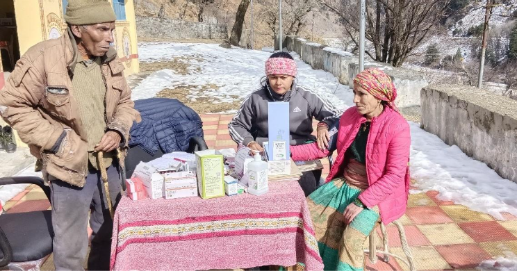 An elderly man stands next to a table with medical supplies, as two women, one wearing a pink jacket and the other a black jacket, interact while seated. The background features snowy terrain and a building.
