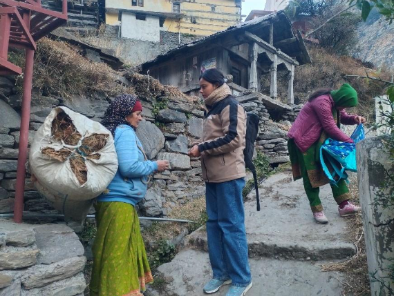 Two women are interacting on a stone pathway in a mountainous area, with one woman carrying a large bag and the other appearing to engage in conversation. A third woman is seen in the background.