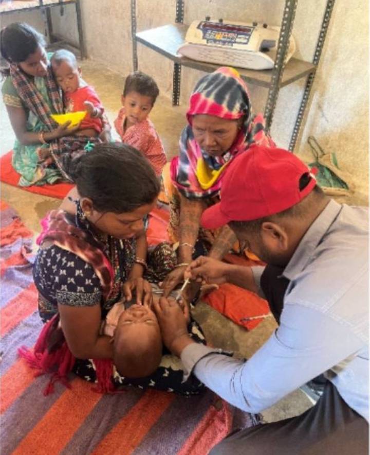 A healthcare worker administering treatment to a baby while surrounded by family members in a communal setting.