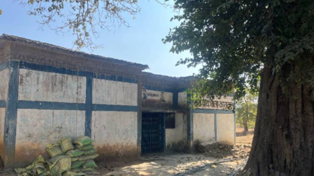 An old building with a blue and white exterior, partially covered in dirt, beside a large tree. Bags of material are stacked near the base of the structure.