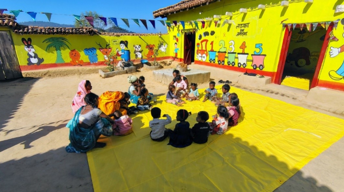 A group of children and adults sitting on a yellow mat in a vibrant outdoor setting with colorful murals on the walls, including animals and numbers. The scene is lively, with children engaging in activities surrounded by nature.