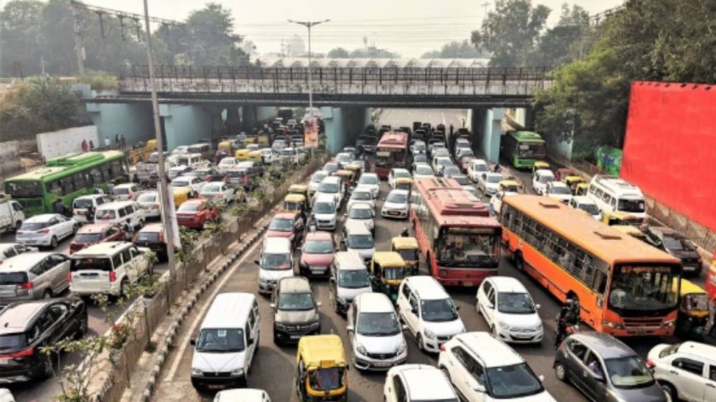 A congested urban traffic scene with numerous vehicles including cars, buses, and auto-rickshaws stuck in heavy traffic under an overpass.