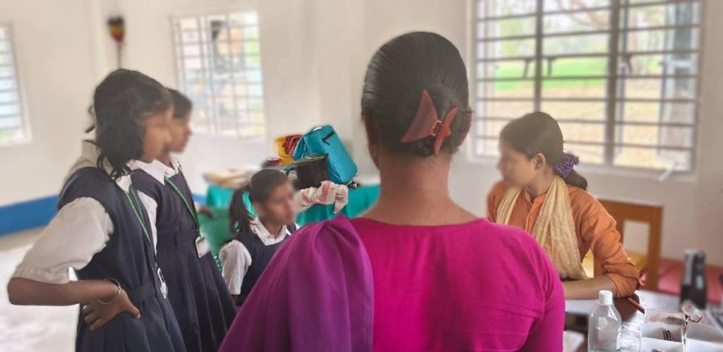 A classroom scene with several schoolgirls in uniforms engaging with a teacher or facilitator, who is wearing a pink top, while another girl in a traditional attire observes.