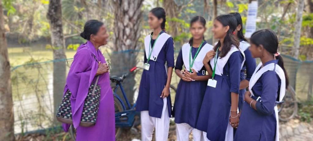An elderly woman in a purple outfit engages in conversation with a group of schoolgirls wearing blue and white uniforms near a rural setting.