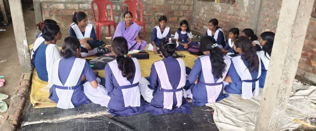 A group of schoolgirls seated in a circle on a mat, engaged in a discussion with a teacher in a casual indoor setting. The walls are made of exposed brick, and a couple of plastic chairs are visible in the background.