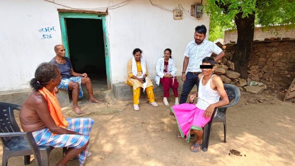 Group of five people sitting outside a house in a rural setting, engaged in conversation. The group includes two older men, a woman in traditional attire, a doctor in a white coat, and a younger man wearing a skirt.