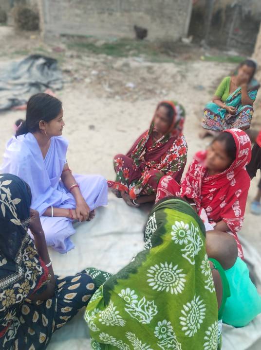 A group of women in colorful traditional attire seated on the ground, engaged in conversation. One woman in a white outfit is speaking to the others, who are wearing vibrant headscarves and dresses.