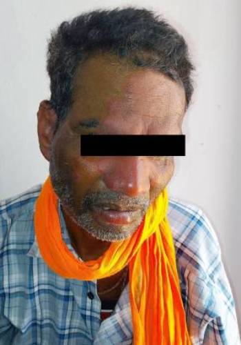 A middle-aged man with short, unkempt hair wearing a blue and white checkered shirt and an orange scarf around his neck, sitting against a plain background.