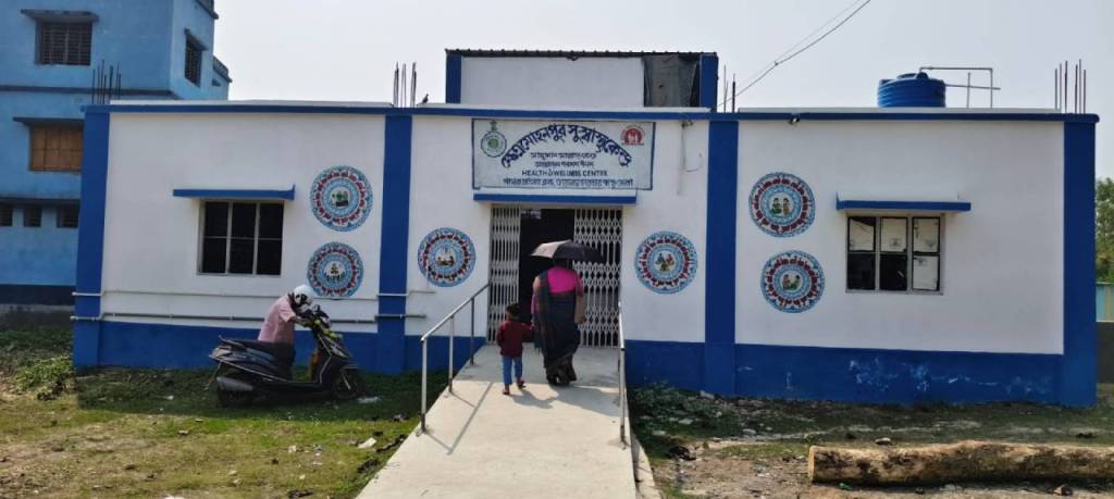 A view of a healthcare center building featuring a blue and white exterior, with several emblems on the walls. Two people, a woman carrying an umbrella and a child, are walking towards the entrance on a cement pathway. A motorcycle is parked nearby.