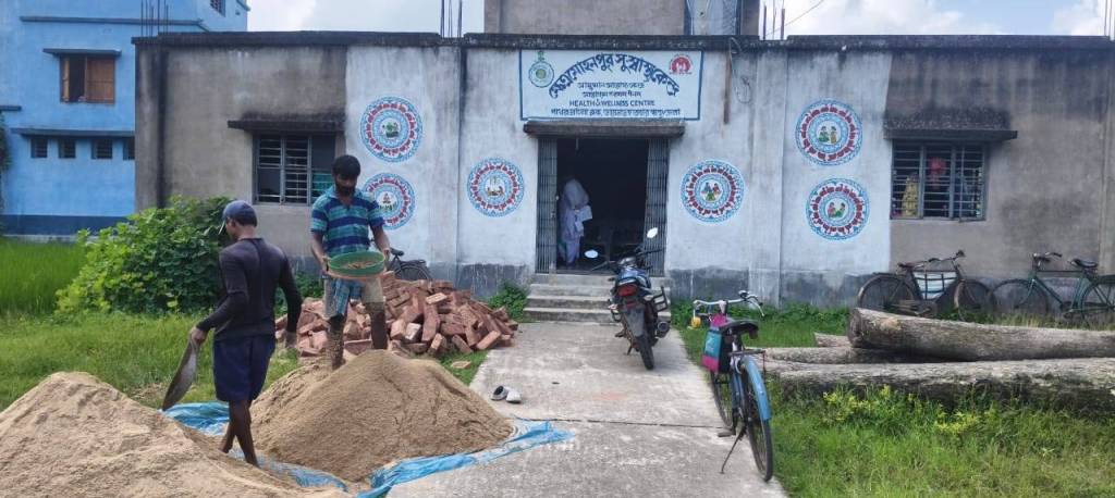 An outdoor scene showcasing two men working with piles of sand and bricks in front of a building labeled as a health and wellness center. Bicycles are parked to the side, and greenery is visible in the background under a partly cloudy sky.