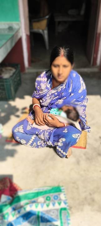 A woman in a blue sari sitting on the ground, holding a baby in her arms, surrounded by a simple outdoor setting.