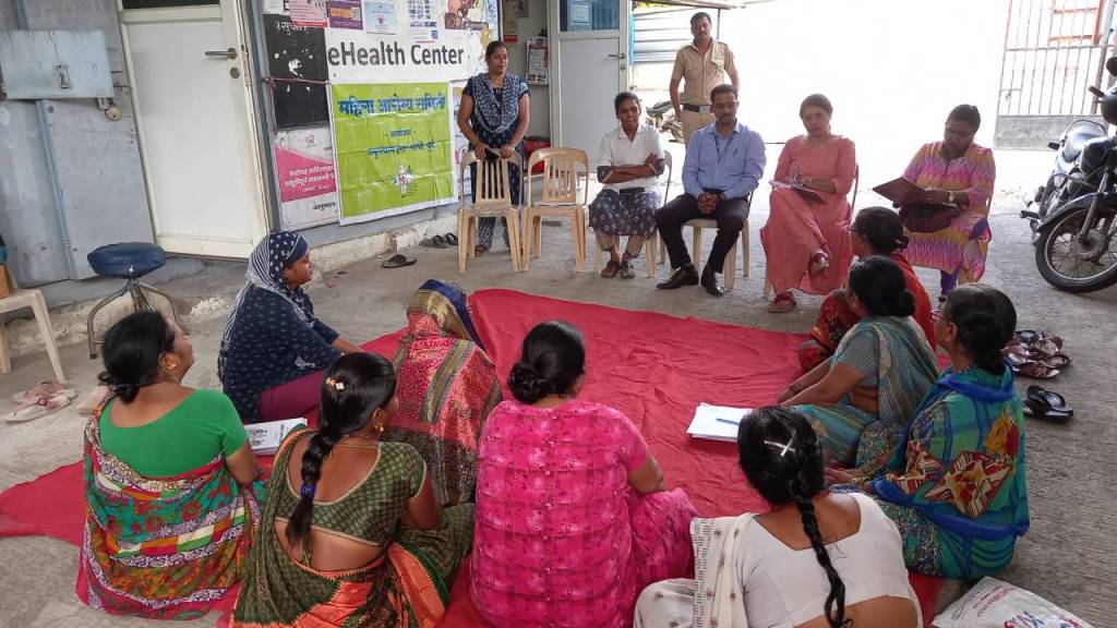 A group of women participating in a community meeting outdoors, seated on a red mat in front of a health center, with some individuals taking notes and others engaged in discussion.