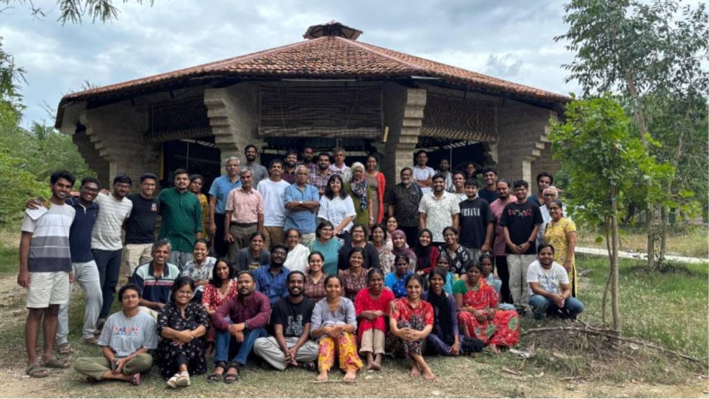 A large group of diverse individuals posing together outside a rustic building with a tiled roof, surrounded by greenery and trees.