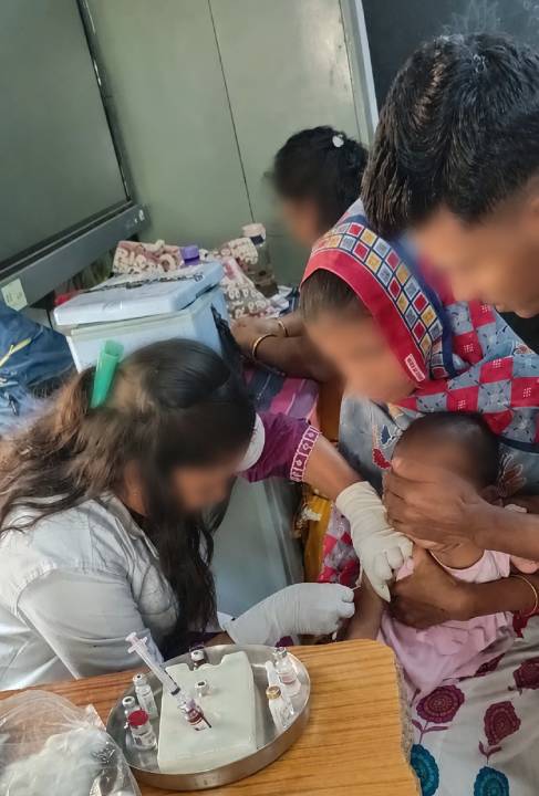 A healthcare worker administers a vaccination to a young child, while a caregiver holds the child. The setting appears to be a clinic or healthcare facility.