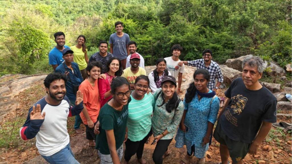 A group of 19 people smiling and posing for a photo outdoors, surrounded by greenery and rocky terrain.