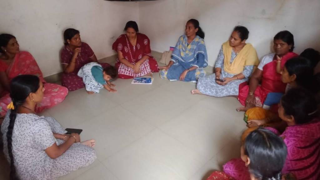 A group of women seated in a circle on the floor, engaged in discussion, with one child playing nearby.